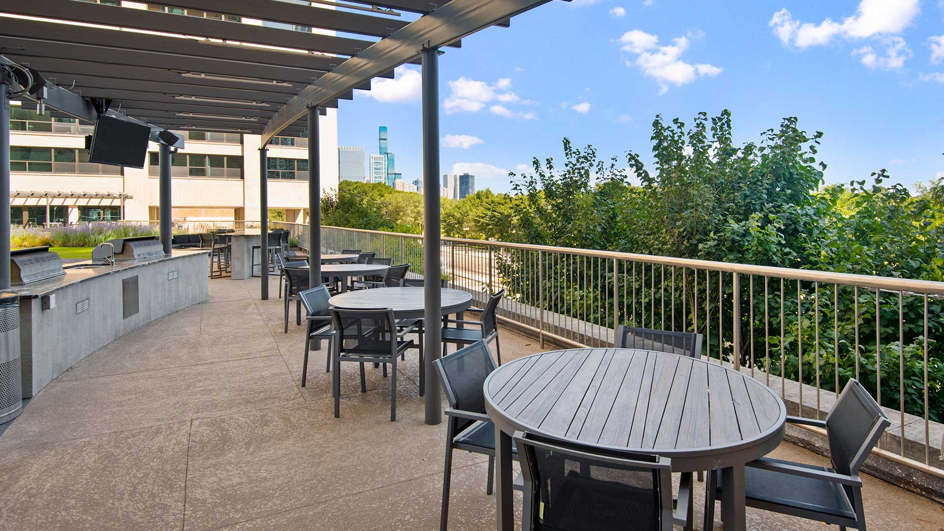Round cafe tables line along the sun deck railing with the grilling stations to the right. Grant Park and the Chicago skyline can be seen in the distance.
