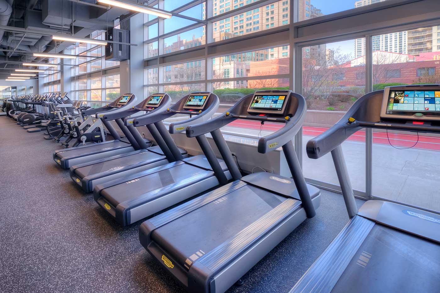 Looking closely at a row of treadmills facing a large window. Outside an outdoor running track is seen, as well as city buildings.