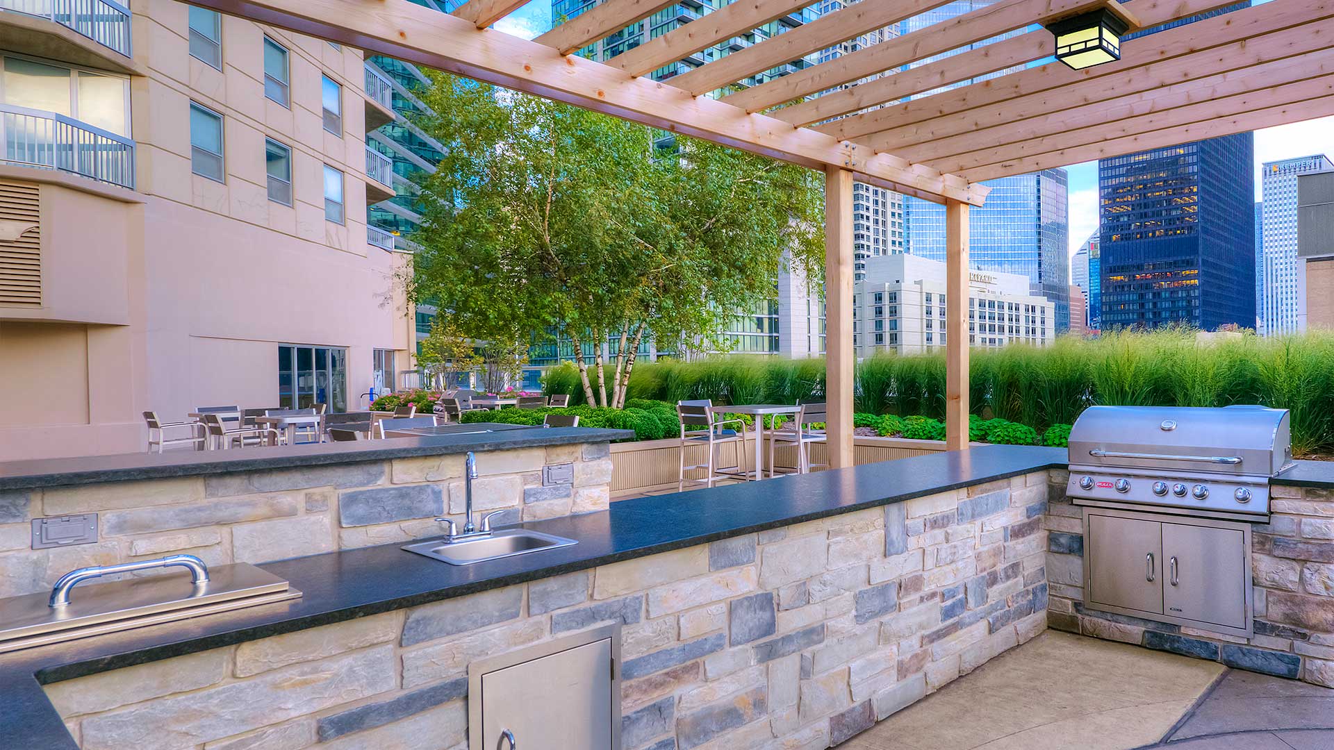 An outdoor kitchen beneath a pergola. The grill is on the right and a sink on the left. Beyond is a seating area and the Grand Plaza apartment tower.