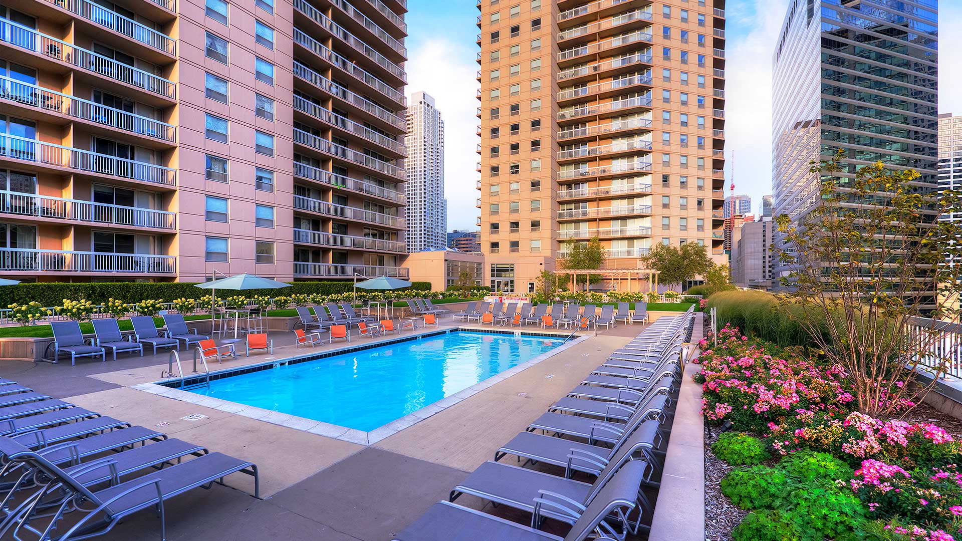 A wide angle shot of the outdoor pool at Grand Plaza. Lounge chairs and café tables surround the pool. There is a row of foliage on the right. The towers of Grand Plaza are seen rising behind and off