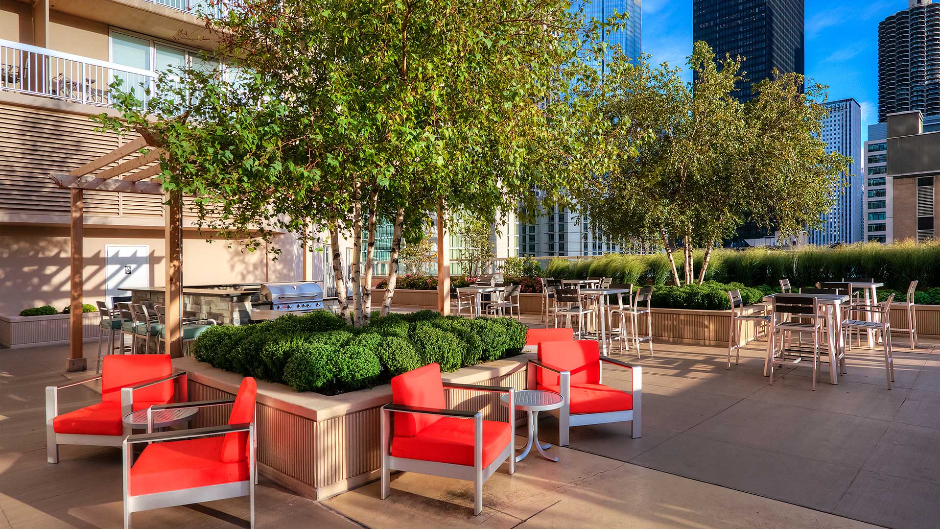 Various outdoor seating areas are set up around two large green areas with trees in the middle. Behind them is a pergola with grilling station.