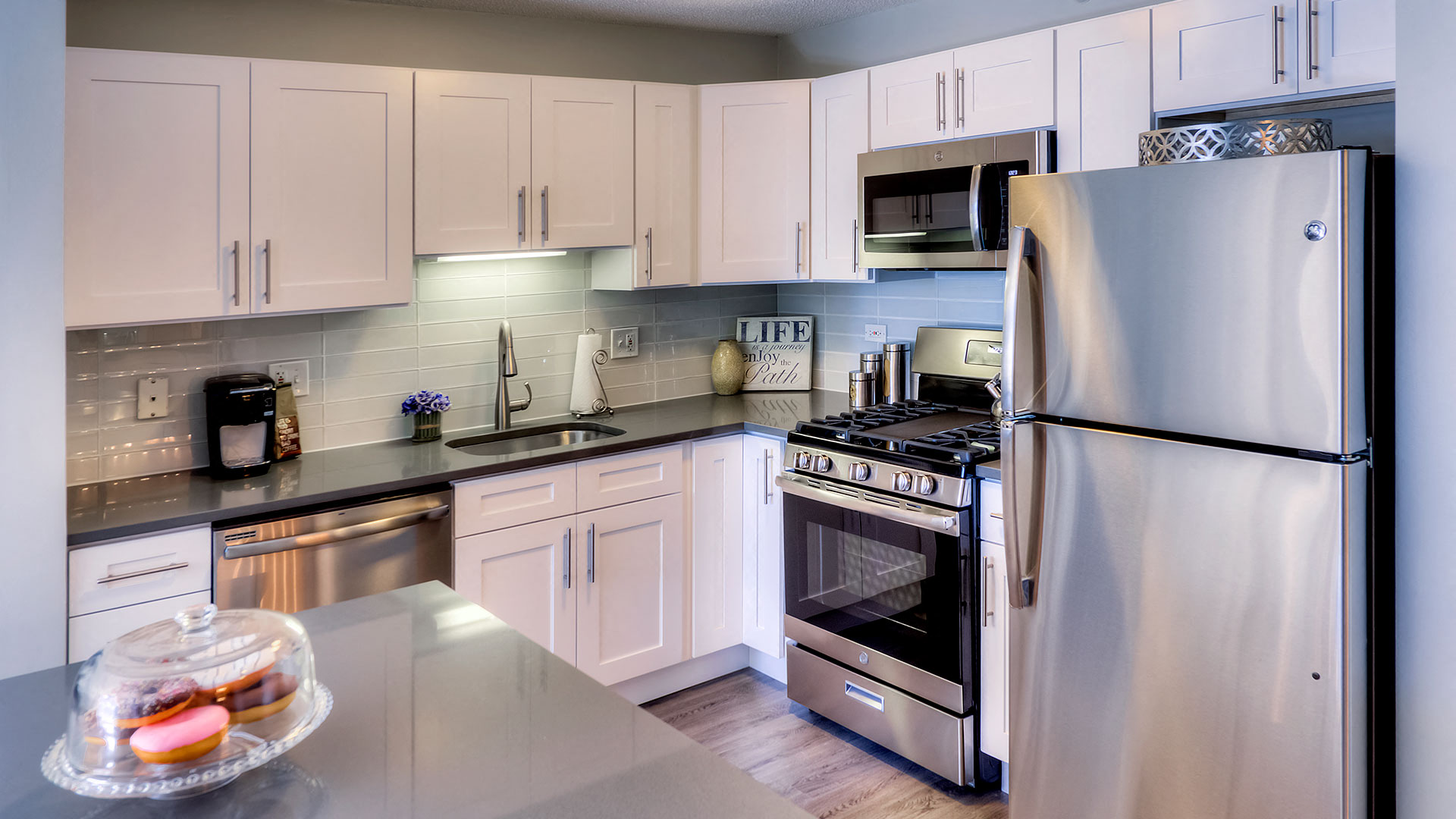 Looking across a kitchen island into the corner kitchen. The refrigerator is on the right with the oven and microwave to the left of it. The kitchen sink is on the left side. The cabinets are white.