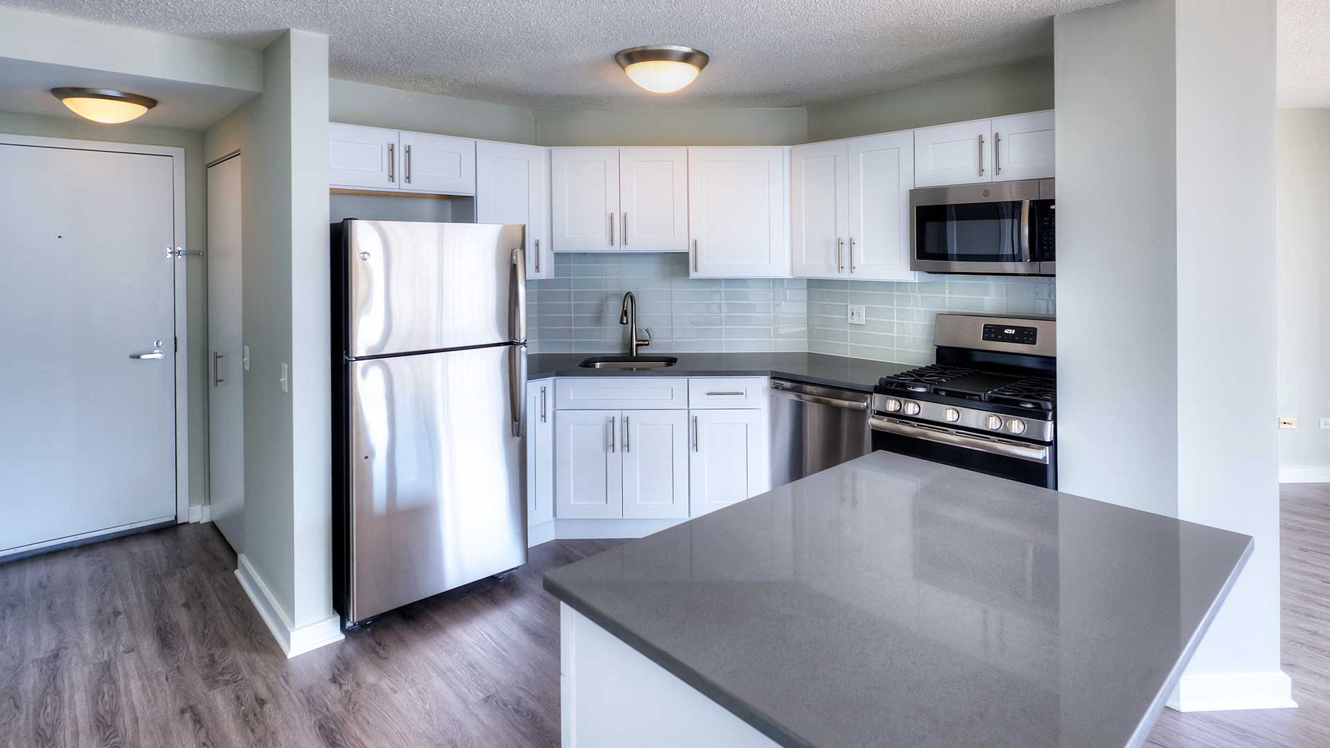 Looking across a kitchen island at a corner kitchen. The stainless refrigerator is on the left, the oven and microwave on the right. The cabinets are white.