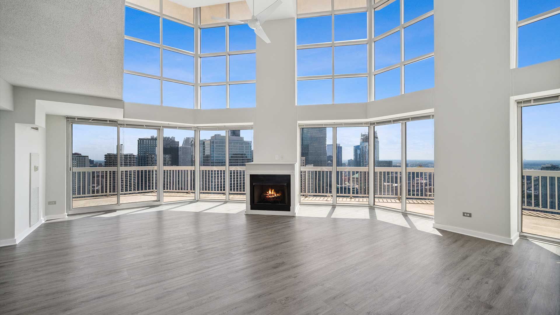 A large open room with windows reaching up two stories. Outside is a balcony and city buildings below. In the center of the room is a column with a lit fireplace at the base.