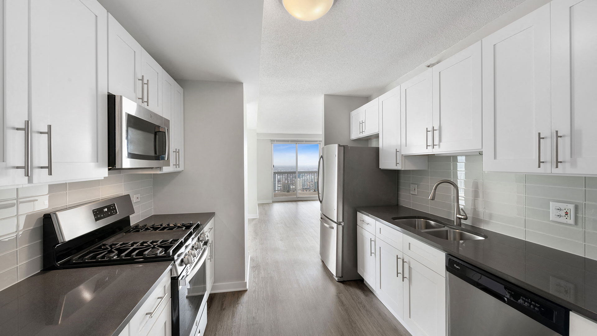 Looking through a galley kitchen into an open area with large glass windows. In the kitchen, the oven and microwave are on the left and the sink and dishwasher are on the right.