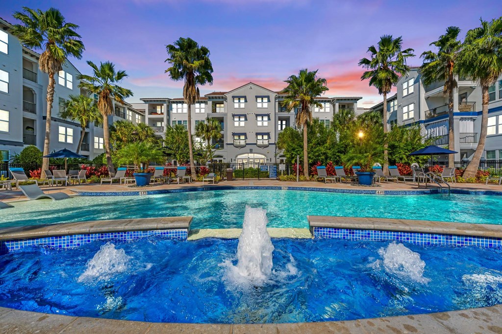 A large swimming pool with a fountain in the middle surrounded by palm trees and apartment buildings.