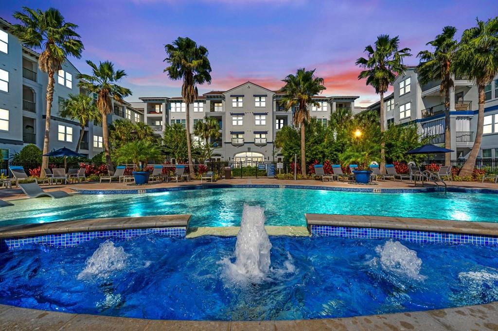 A large swimming pool with a fountain in the middle surrounded by palm trees and apartment buildings.