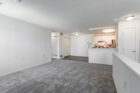 the living room and kitchen of an apartment with white walls and carpet