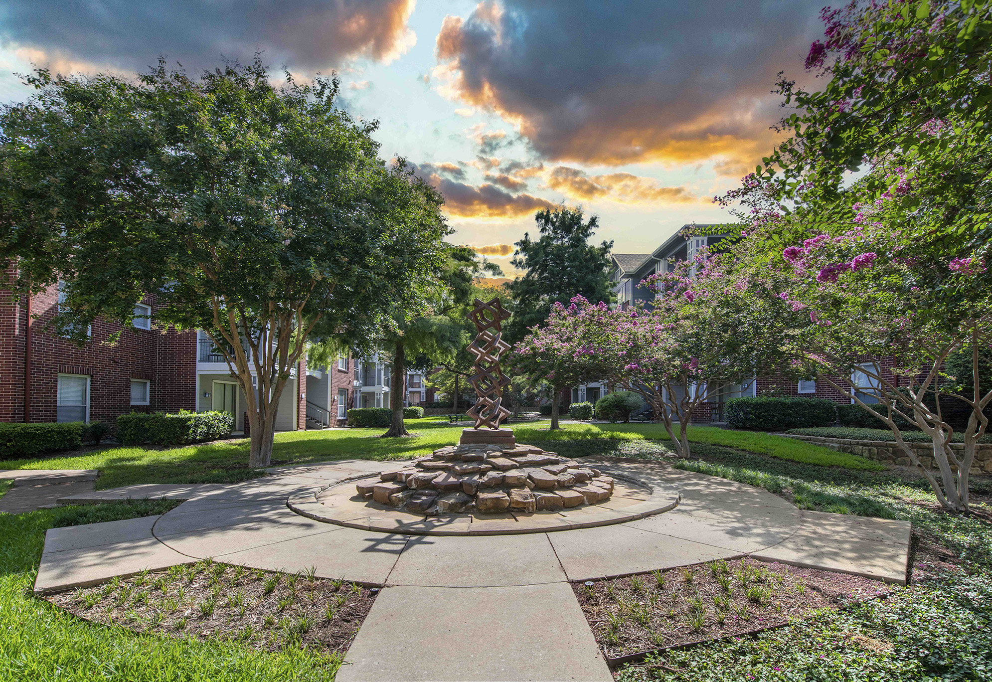 a fountain in the middle of a park with trees and a sunset