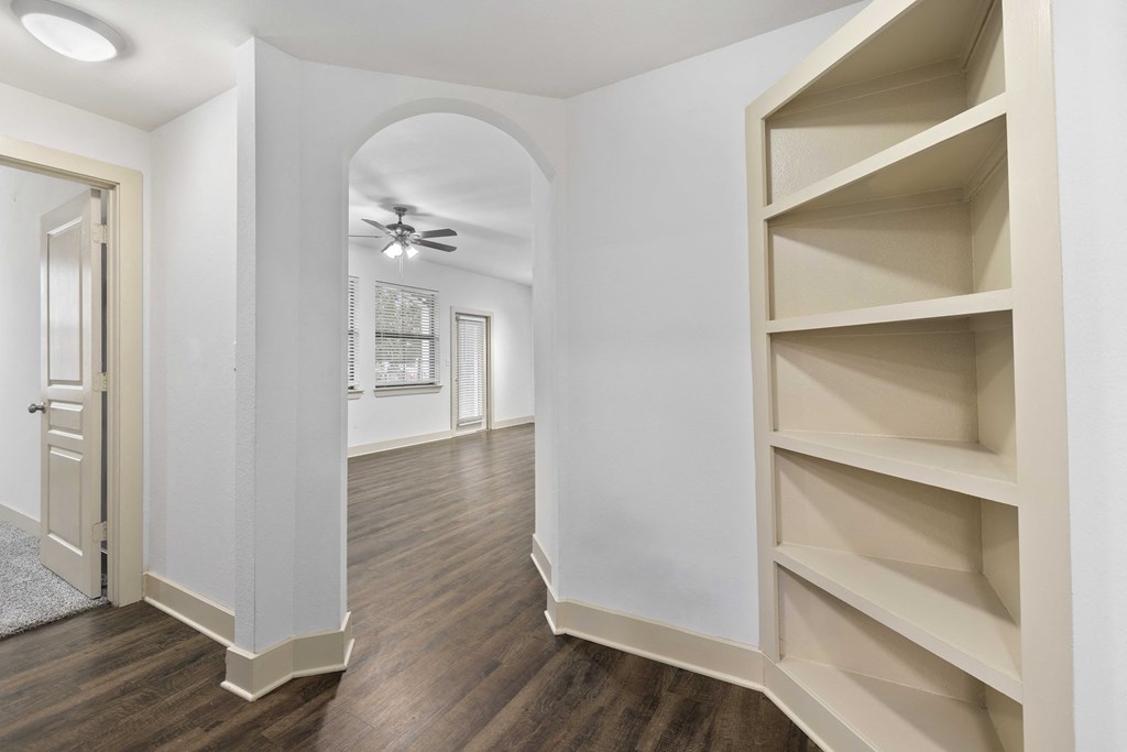 A room with a wooden floor and a white wall with a shelf on the right side.
