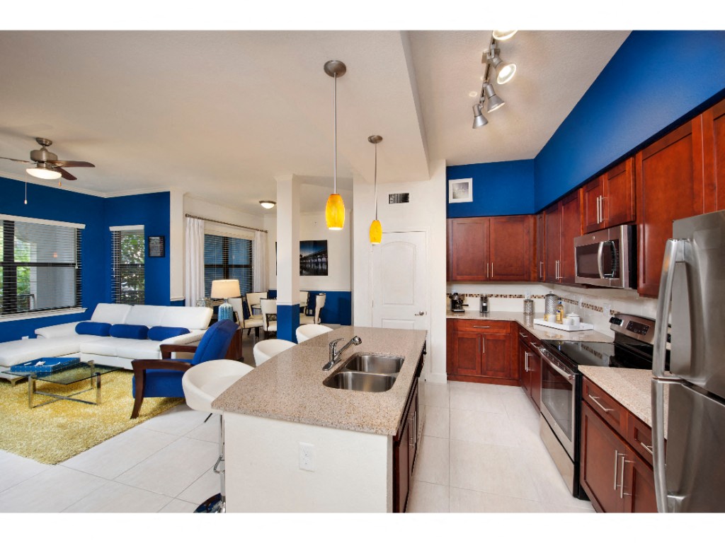 Kitchen with sink in island, stainless steel microwave, oven, and refrigerator next to living room with couch, chair, coffee table, and ceiling fan