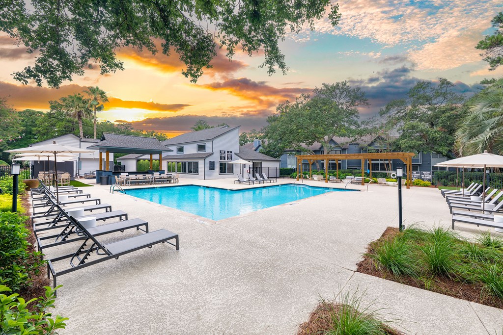 A pool area with sun loungers and a building in the background during sunset.