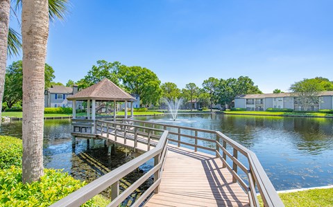 a wooden bridge leading to a lake with a gazebo and water fountain