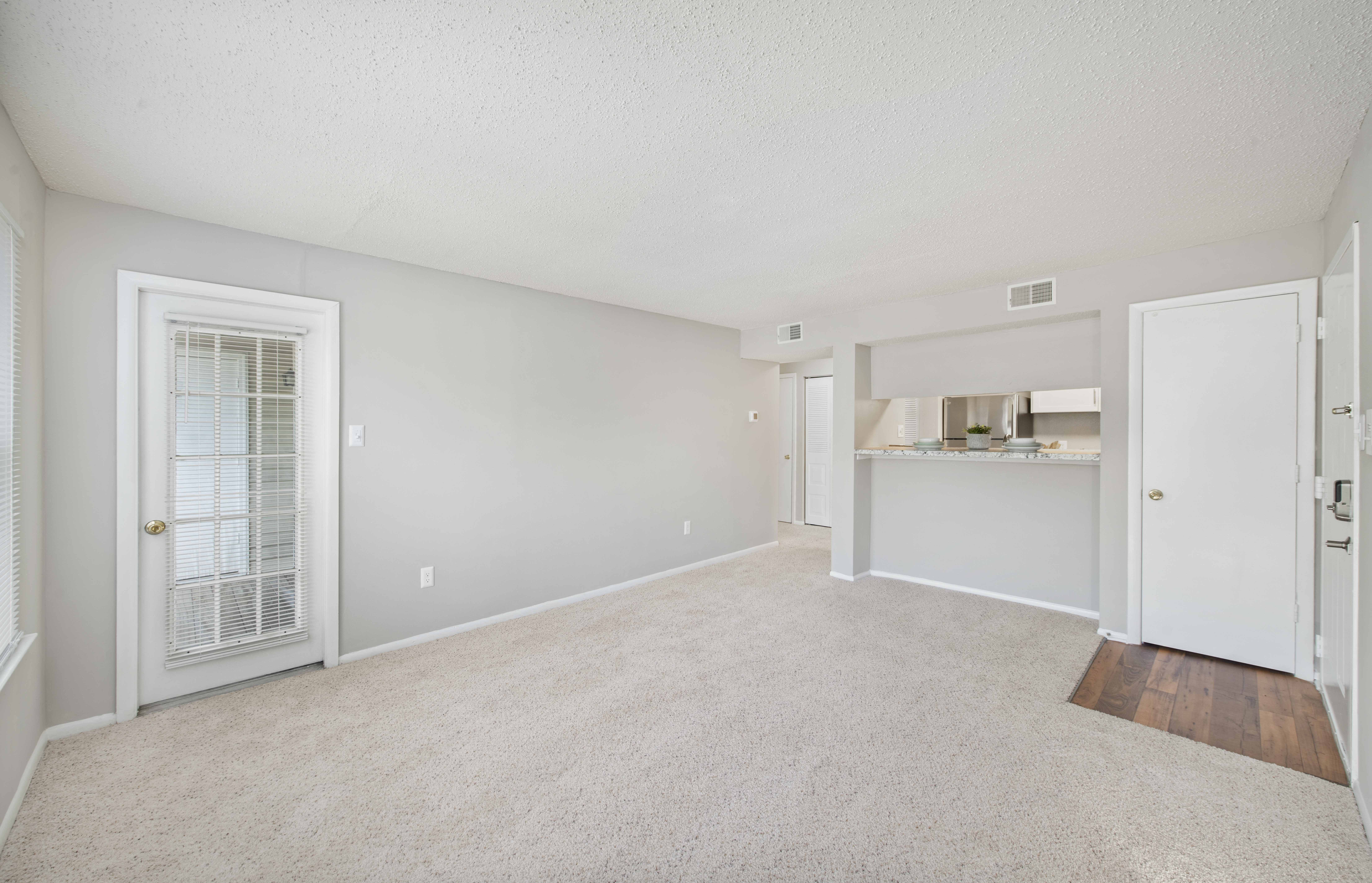 the living room and kitchen of an apartment with white walls and carpet