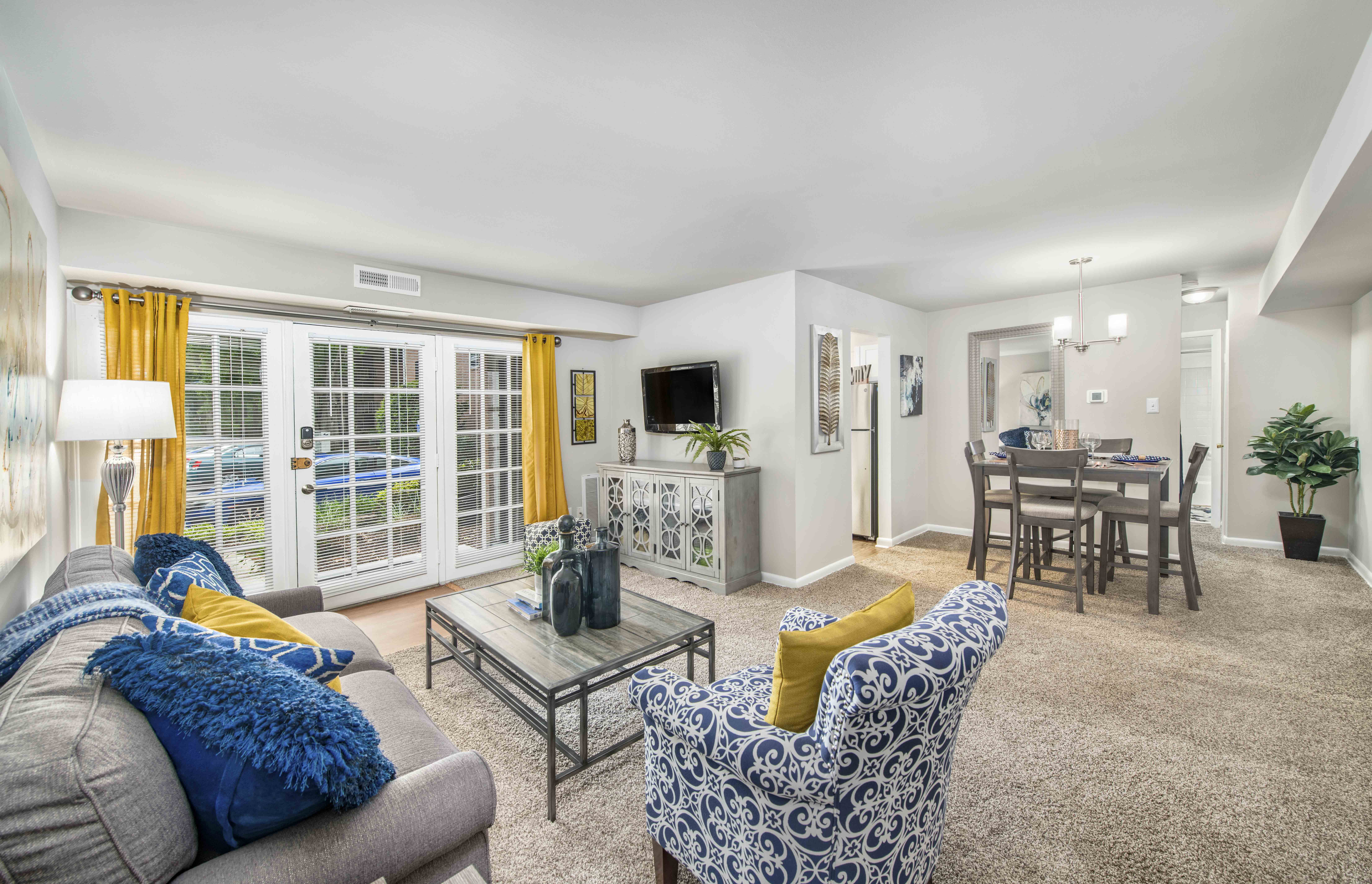 A living room with a grey couch, a blue and white patterned armchair, a glass coffee table, and a dining area with a table and chairs.