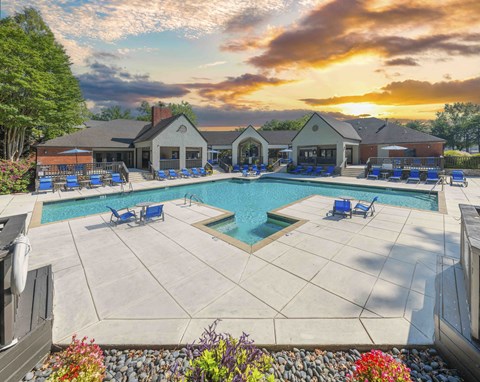 A large outdoor swimming pool surrounded by a patio area with chairs and a building in the background.