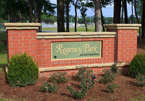 A sign for Regency Park Apartments is framed by a brick wall and bordered by shrubbery.