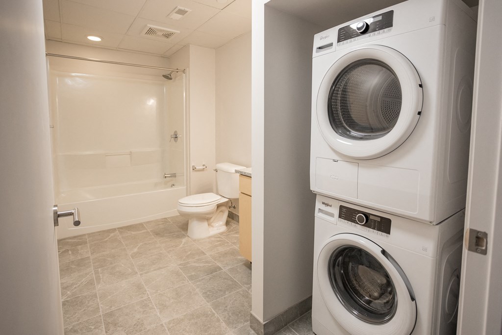 A white washing machine is in a small laundry room with a toilet and a bathtub.