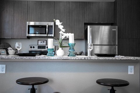 A kitchen with a stainless steel refrigerator and a microwave above a counter with two black stools.