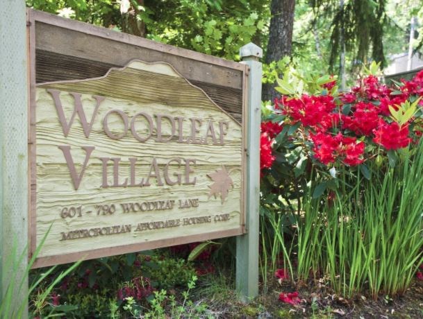 A wooden sign that says Woodear Village in front of some flowers.