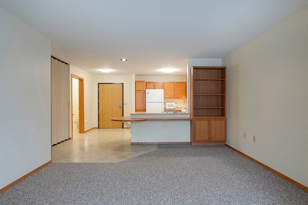 an empty living room with a kitchen and a door to a hallway