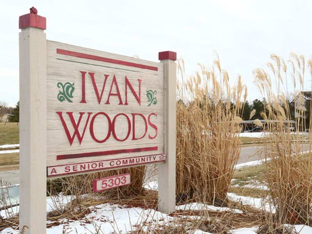 Sign for Ivan Woods, a senior community, with red lettering on a white wooden board, surrounded by tall dried grasses and light snow on the ground, with a suburban backdrop