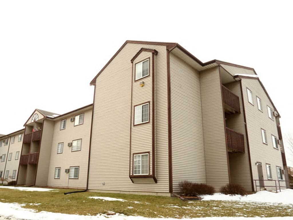 Three-story beige apartment building with brown trim, featuring balconies and multiple windows, set in a winter landscape with patches of snow on the ground and an overcast sky