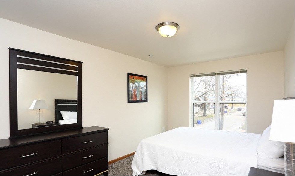 A bedroom with a white bed, a black dresser with a mirror, and a window with a view of trees.
