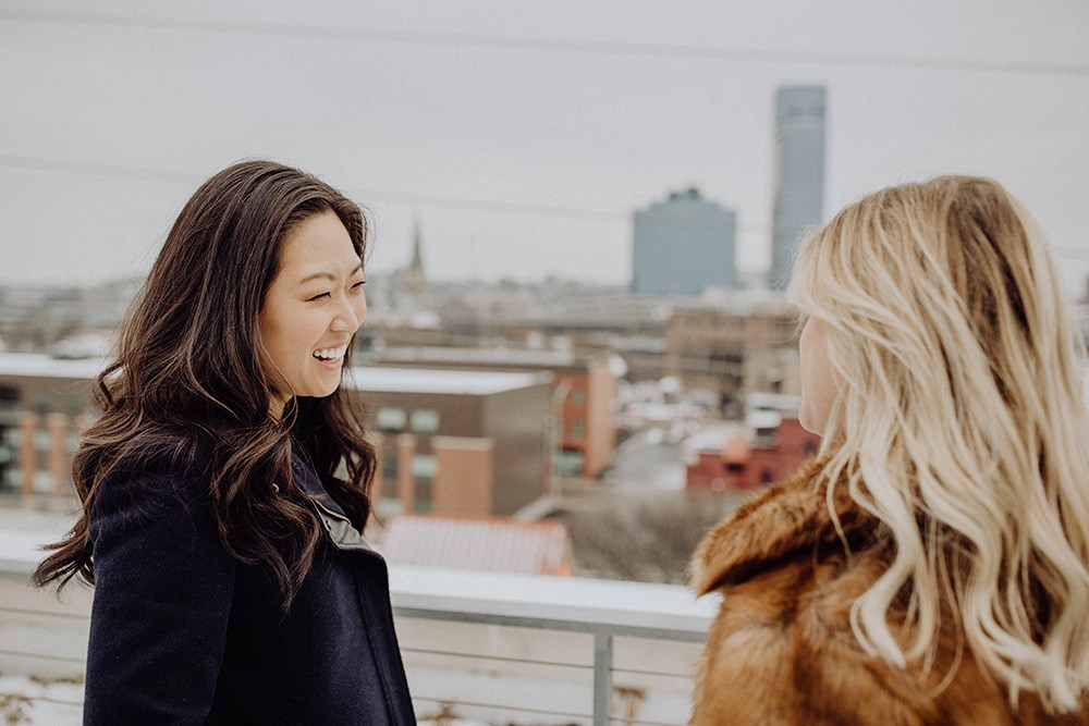 two women laughing on a rooftop with a city in the background