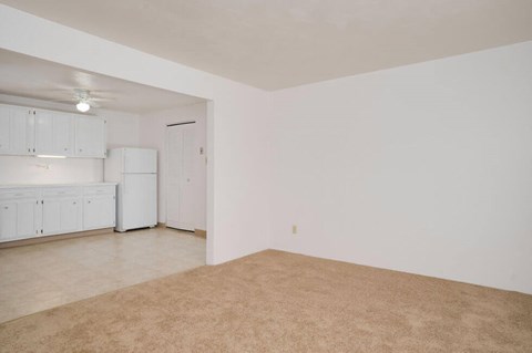 the living room and kitchen of an apartment with white walls and carpet