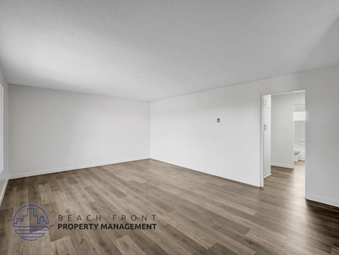 A spacious room with wooden flooring and white walls, with a logo for Beach Front Property Management in the bottom left corner.