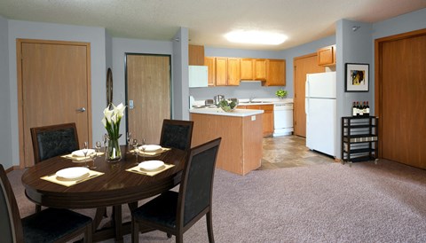 A dining room with a table set for two and a kitchen in the background.