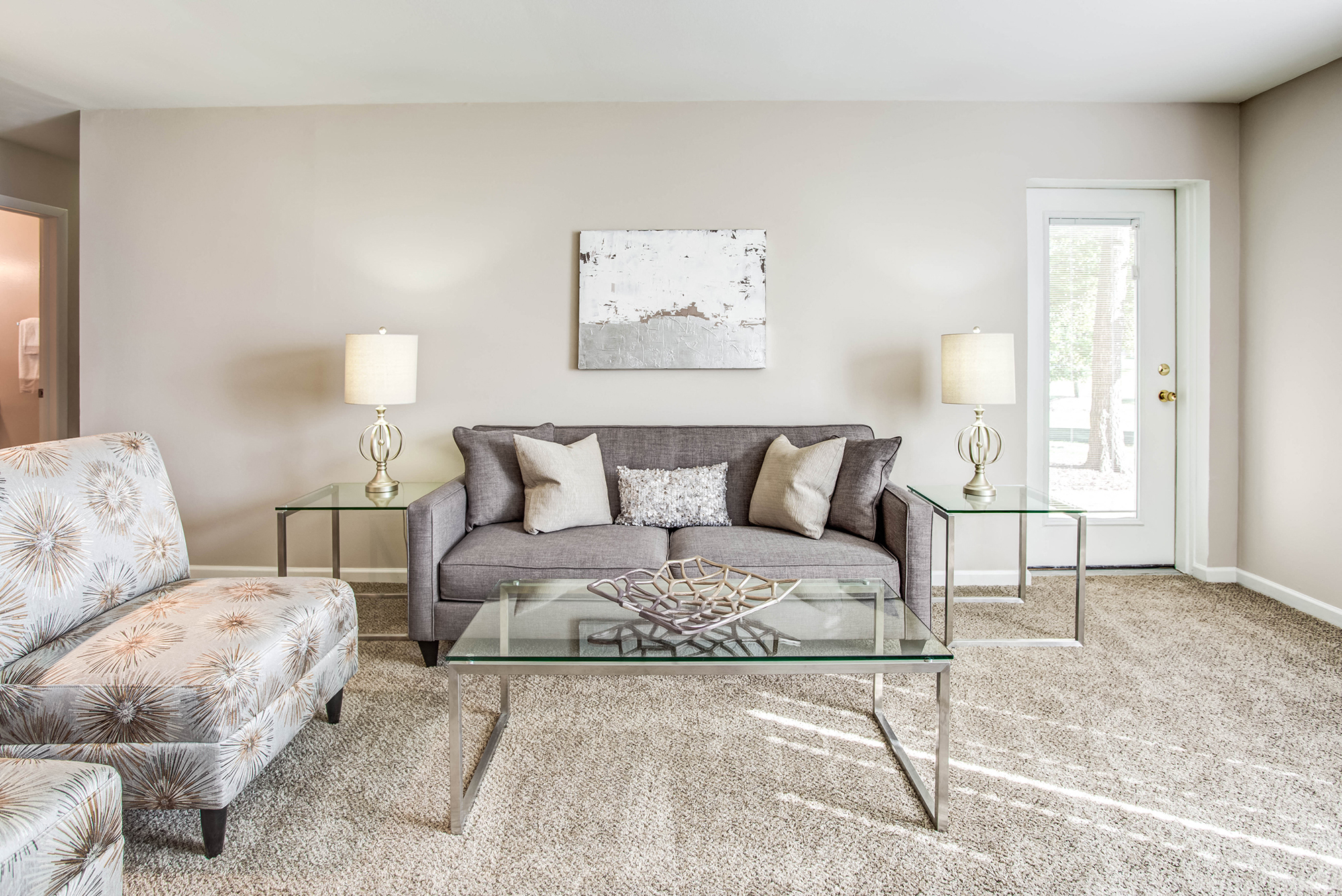 A living room with a grey couch, a glass coffee table, and a painting on the wall.