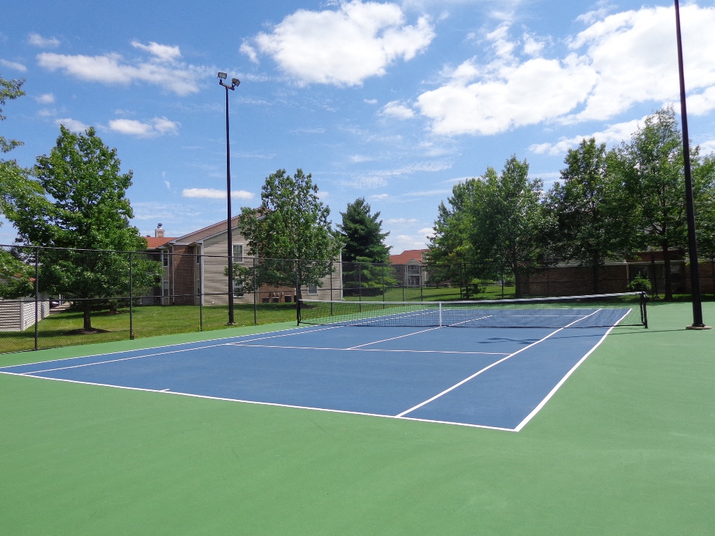 a blue and green tennis court with trees and buildings in the background