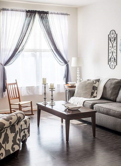 A living room with a grey couch, a wooden chair, a patterned ottoman, a table with a bowl and a candle on it, and a window with curtains.