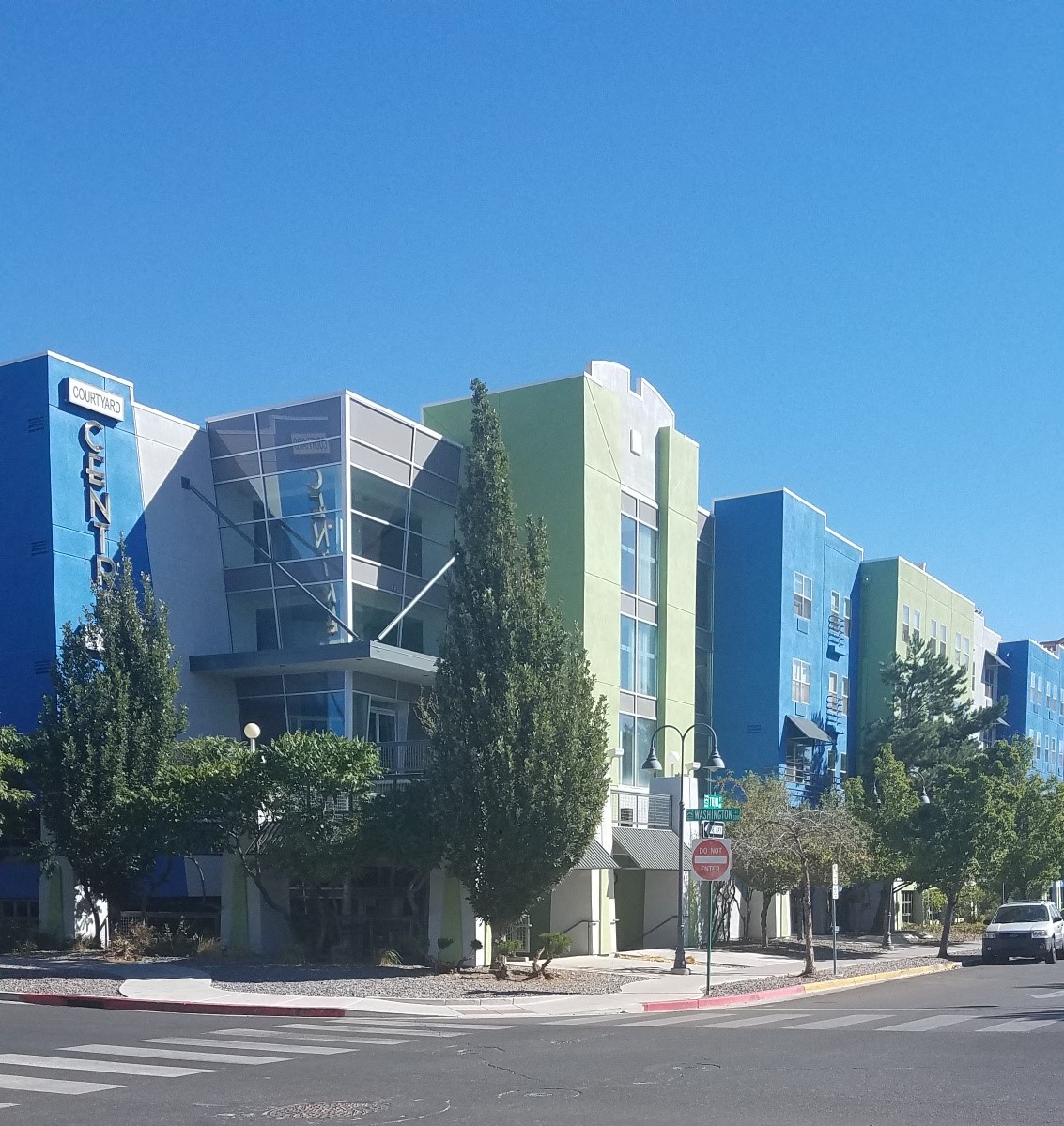 A modern building with a blue and green facade is surrounded by trees and has a clear blue sky in the background.