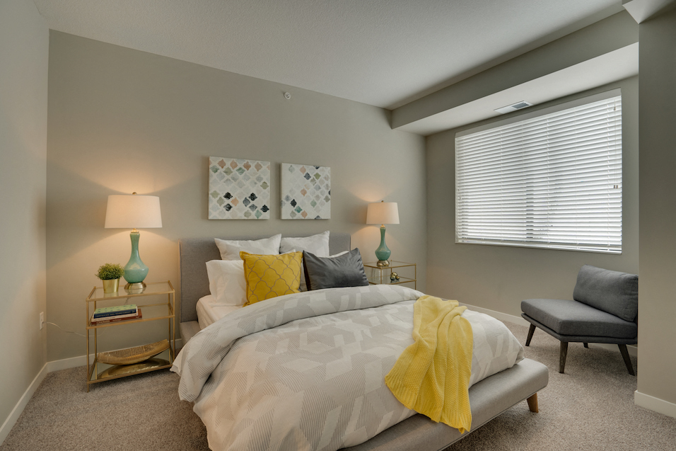 Bed with white comforter and different colored pillows, endtables with lamps, and a gray chair in the corner by the window