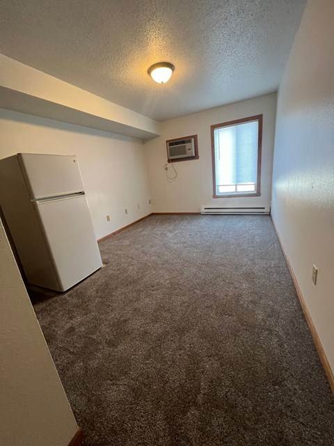 A room with a carpeted floor, a window with blinds, and a white refrigerator.