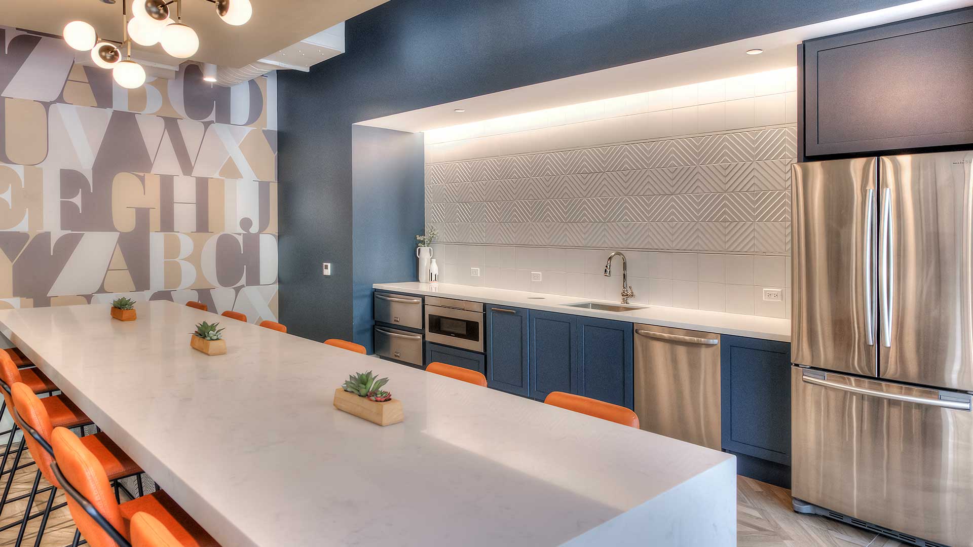 Looking across the coffee bar at an angle in the lounge at Burnham Pointe. There are orange stools set on both sides of the bar. The back wall has built-in appliances and there is a large refrigerator