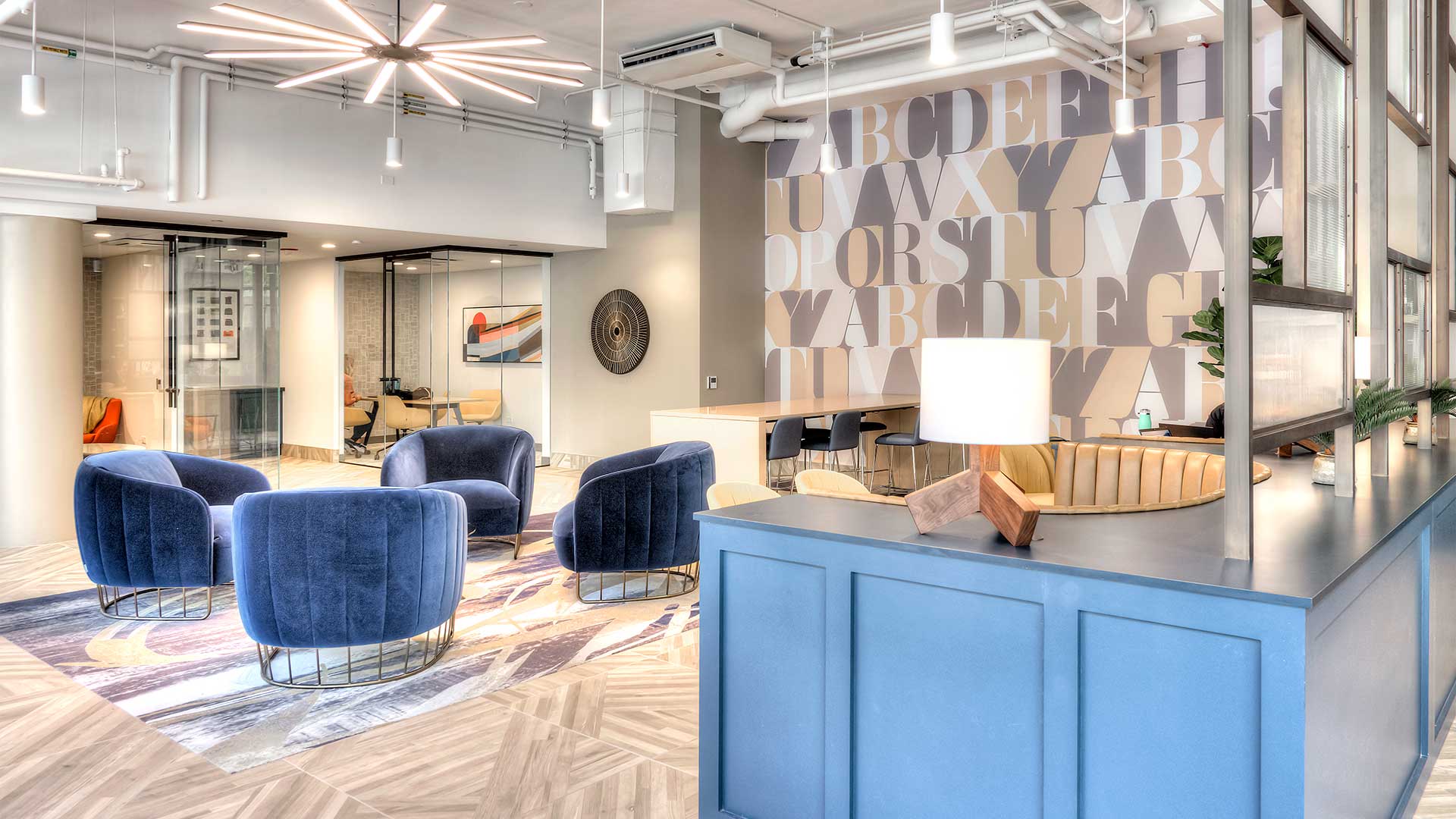 Looking towards the private co-working spaces from the entertainment lounge. The co-working spaces are behind glass walls at the back. There are various club chairs and booths for seating.