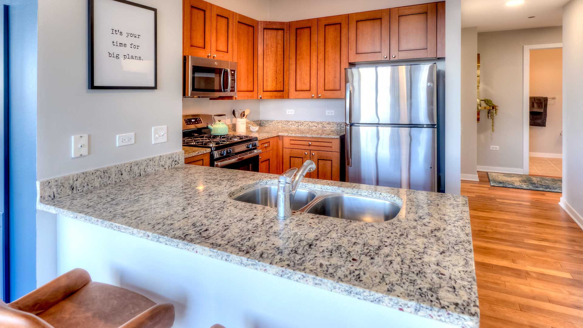 Looking across a kitchen counter in a residence in Burnham Pointe. There is an undermount sink on the counter. Behind is the rest of the kitchen with brown cabinets, an oven and microwave to the left