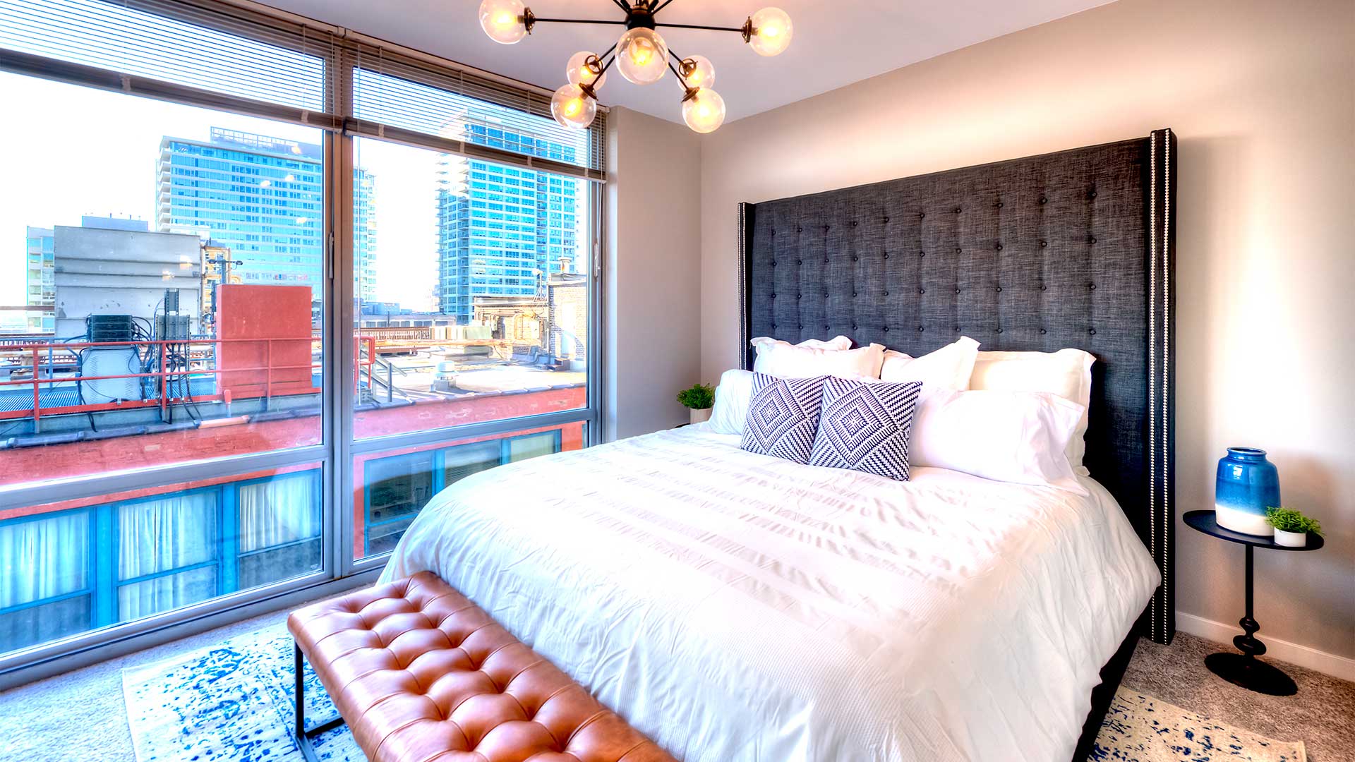 A large bed with a bench and the foot sits before large, floor-to-ceiling windows that show the city outside.