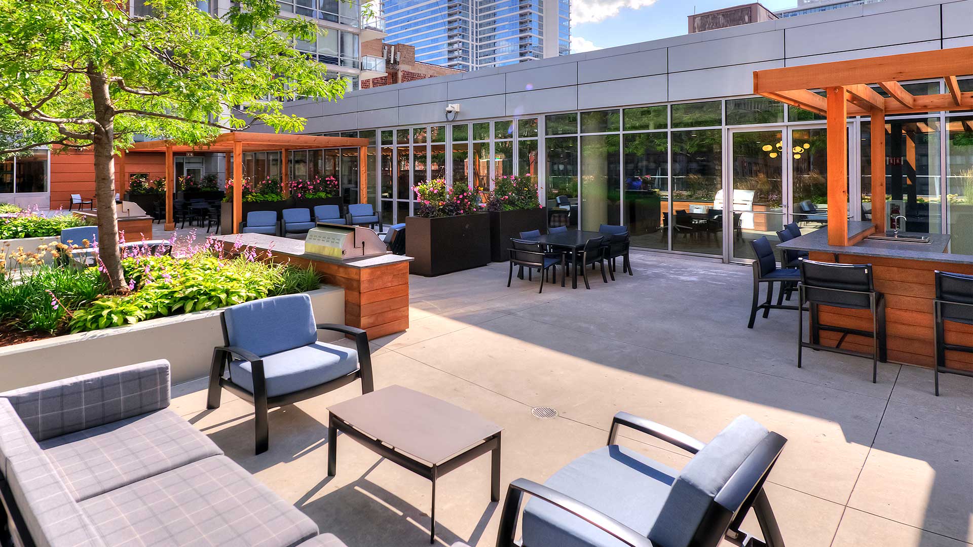 An outdoor furniture set sits beside potted plants with more seating further back. The grilling area is back and to the right.
