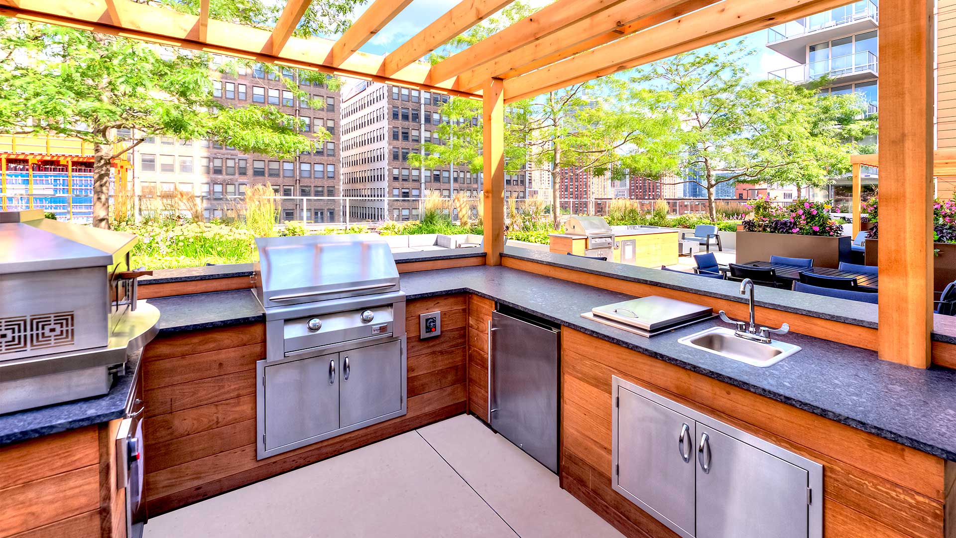An outdoor kitchen with built-in grill to the left and an under-counter fridge and sink to the right.