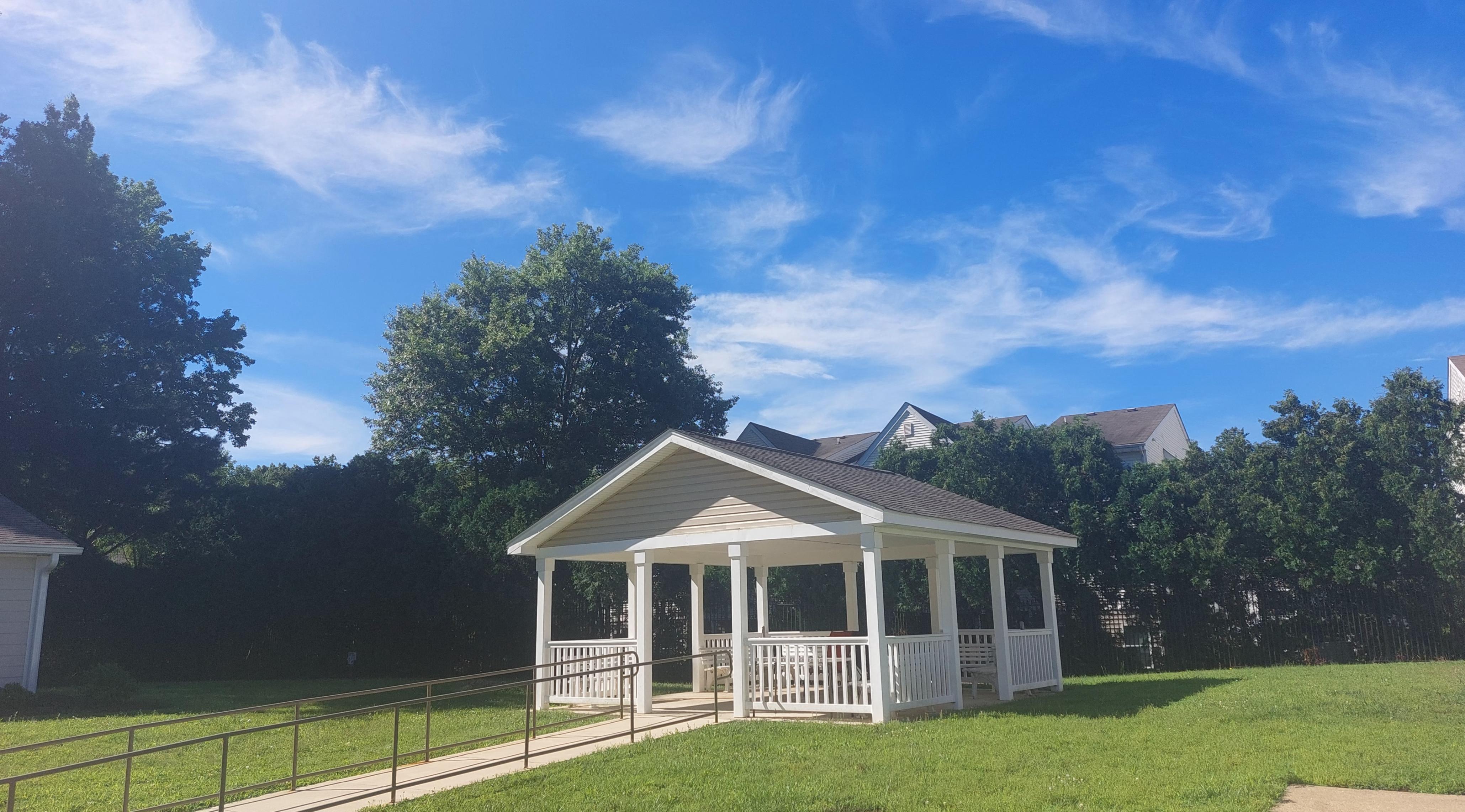 A small white gazebo with a black roof is surrounded by a grassy area and trees.