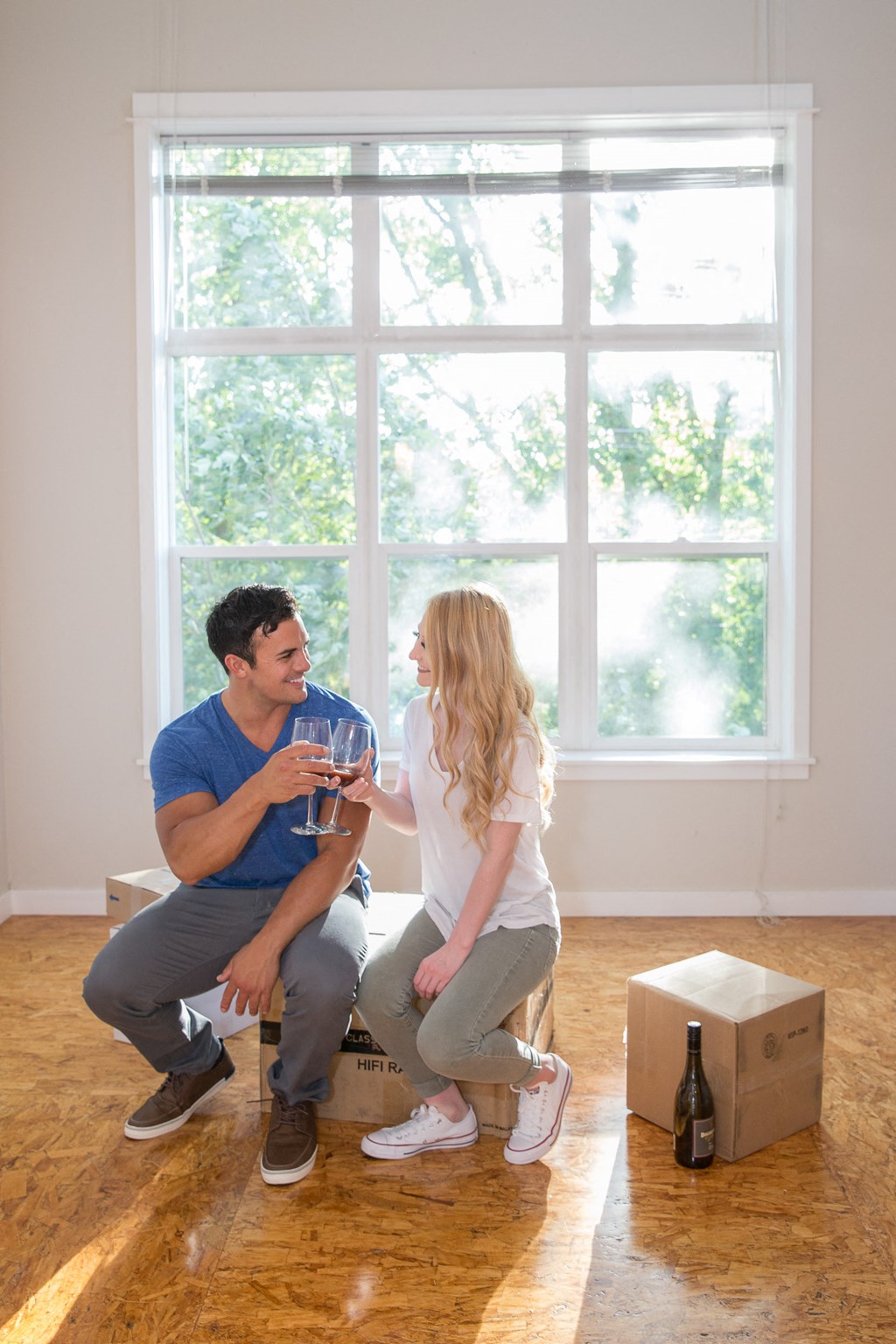 a man and woman sitting on a window sill in an empty room drinking wine