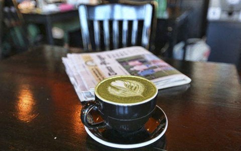 A cup of coffee with latte art on top sits on a saucer with a newspaper and a chair in the background.