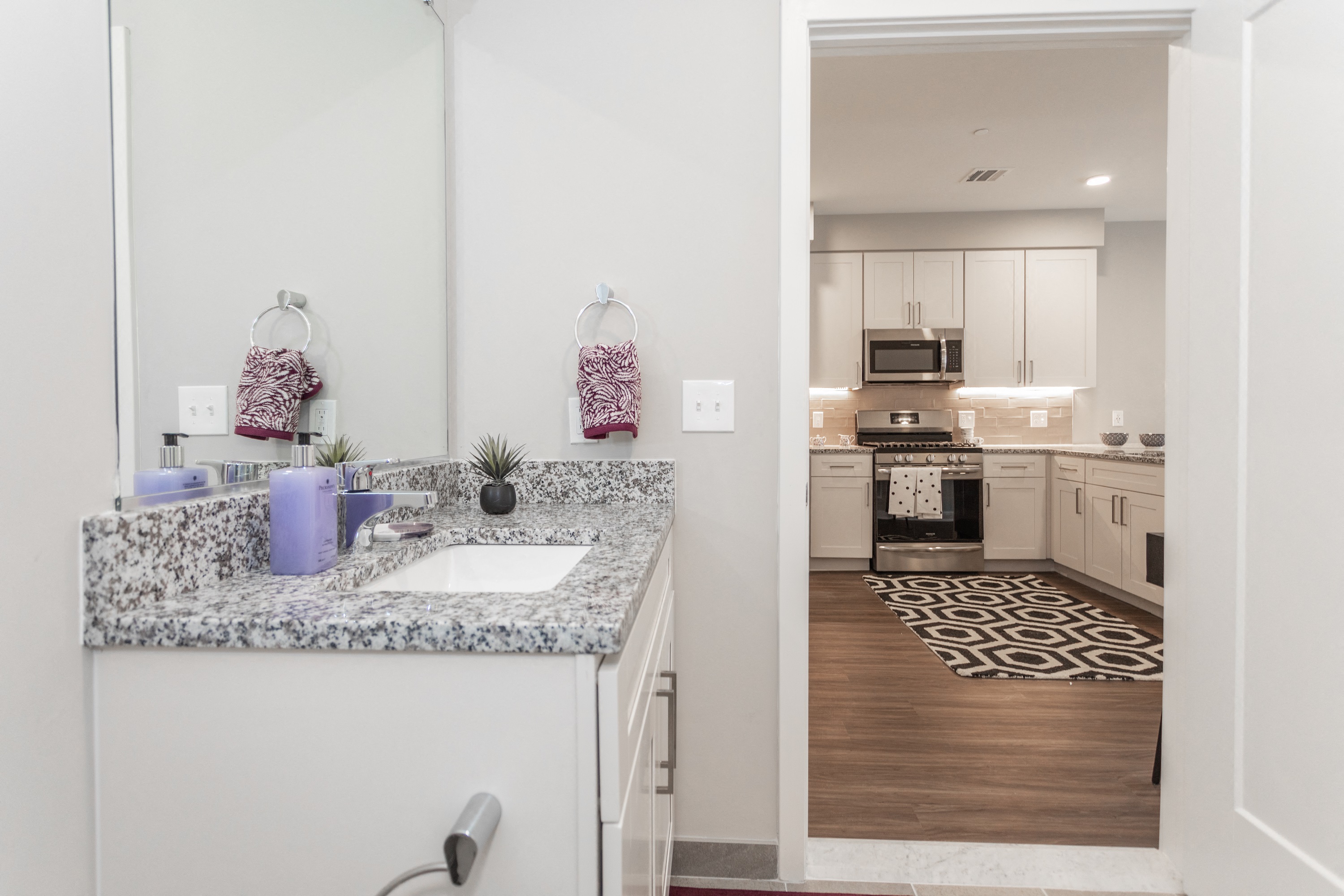 Apartment bathroom with marbled countertop, white vanity, and view in to kitchen