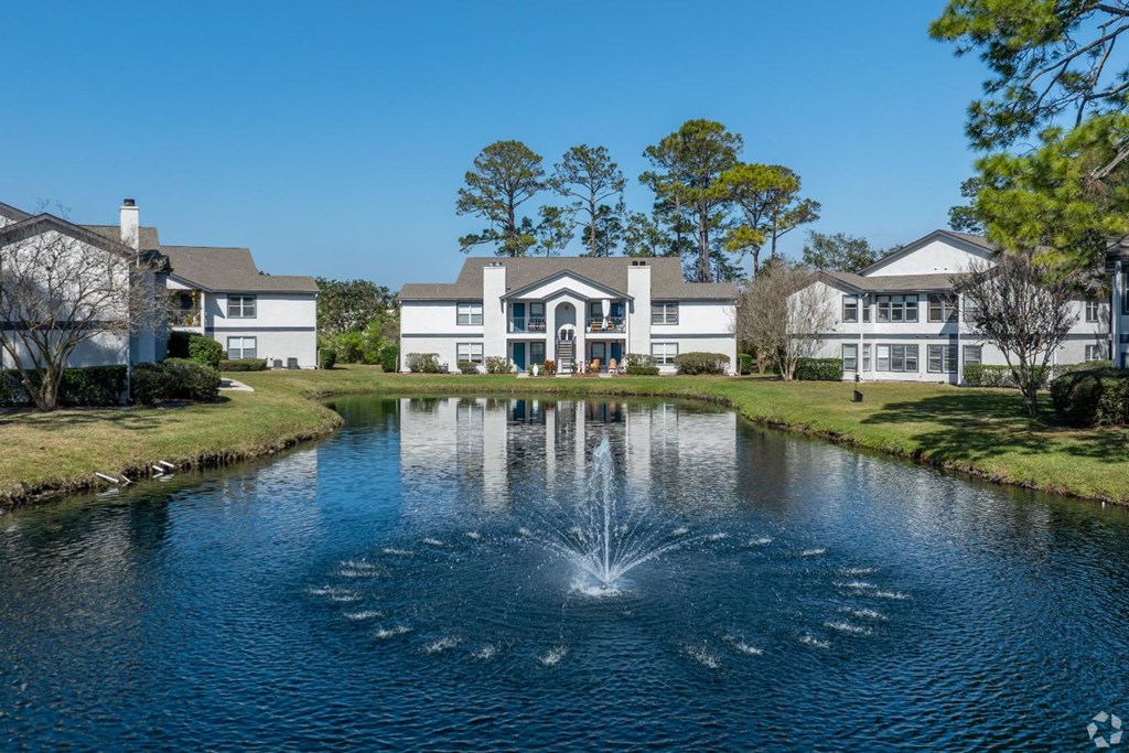 A fountain in the middle of a pond in front of a row of houses.