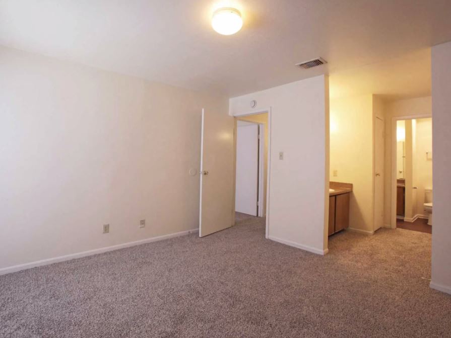 Spacious bedroom with sandy colored carpets and eggshell white walls with partial view of bathroom